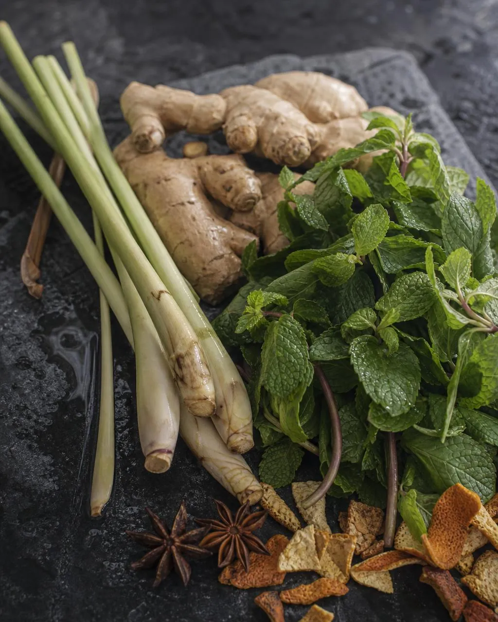 Fresh Thai botanicals — lemongrass, mint, ginger, orange peel and anise — on dark stone